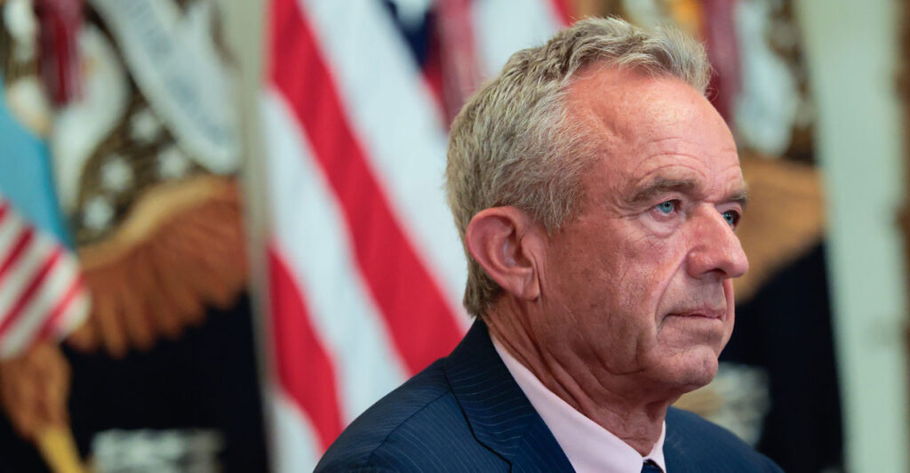 Robert F. Kennedy Jr. sits at a desk during a news conference and stares straight ahead.