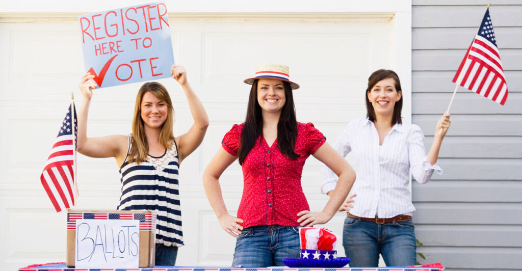 Three young women staff a voter-registration table, with a mockup of a ballot box and two small U.S. flags.