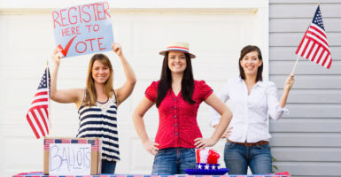 Three young women staff a voter-registration table, with a mockup of a ballot box and two small U.S. flags.