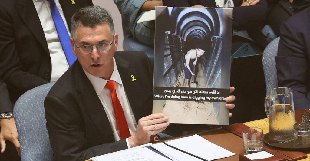 Gideon Sa'ar sits at a table and holds up an enlarged photo of Israeli captive Evyatar David while speaking.
