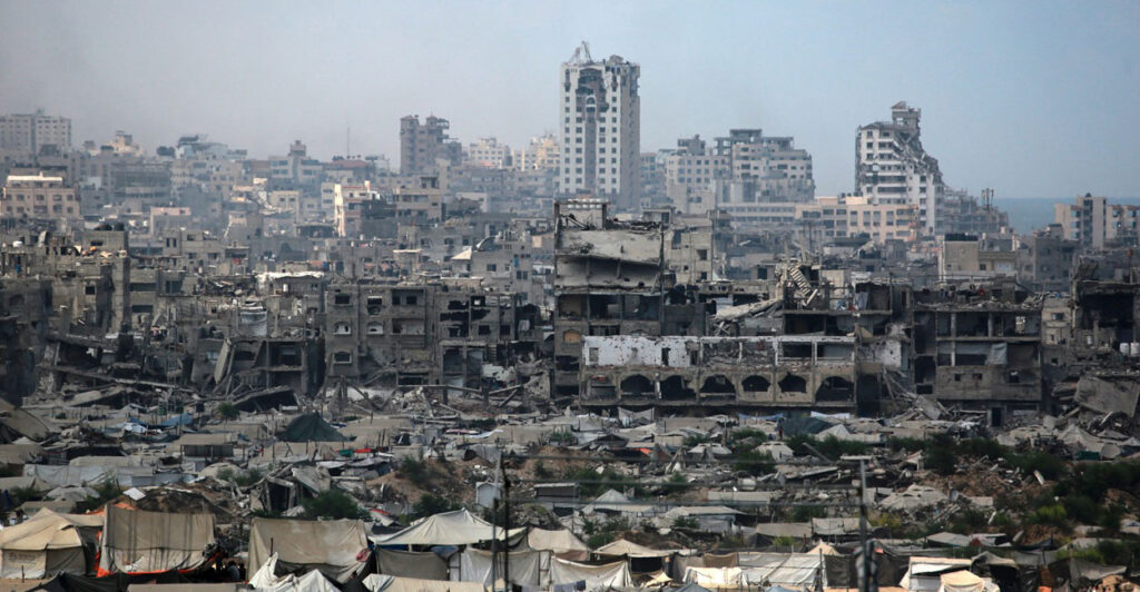 The war torn skyline of Gaza City is shown with tents set up beneath crumbling buildings.