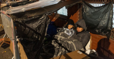 Migrants sit outside in a covered area that has a plastic lining attached to a rail to form a tent.