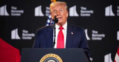 Donald Trump stands in front of a podium and speaks into a microphone with his hands at his side.