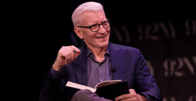 Anderson Cooper sits with his legs crossed as he holds a book in his lap with his left hand.