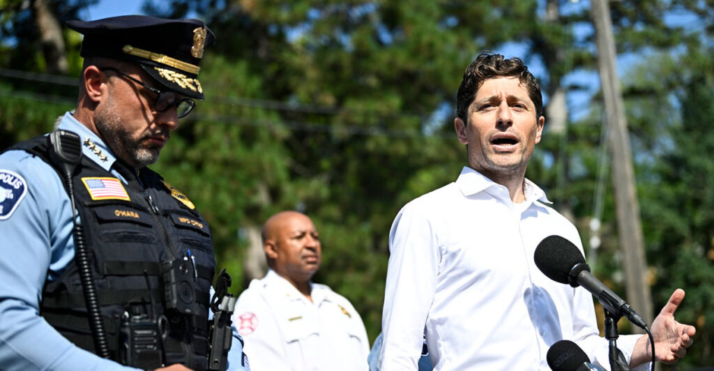 Jacob Frey stands outdoors in front of a microphone and speaks to the media while gesturing with his left hand.