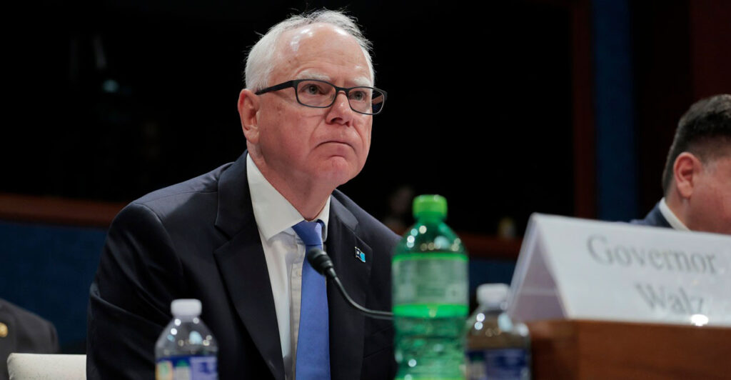 Tim Walz sits at a table near a plastic bottle and a microphone as he listens during a hearing.