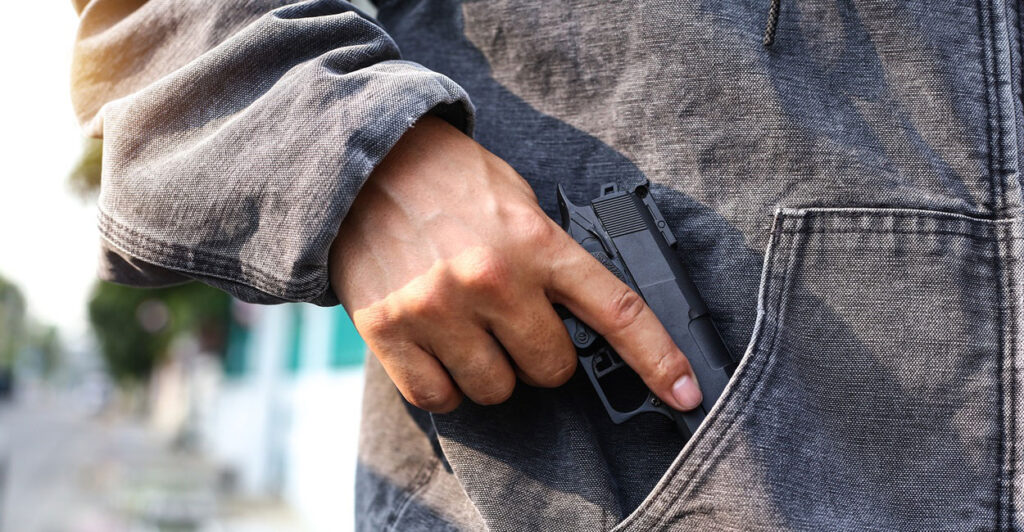 A person wearing a jacket holds onto a gun with their right hand and has it partially hidden in a pocket.