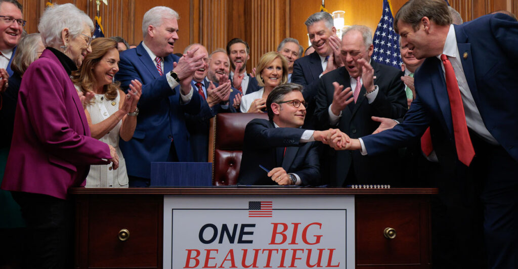 Speaker Mike Johnson shakes hands at a table with a banner promoting the "One Big Beautiful Bill Act."