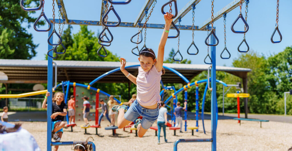 High-flying young girls swings with one arm from a jungle-gym as another girl looks on.