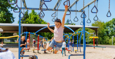 High-flying young girls swings with one arm from a jungle-gym as another girl looks on.