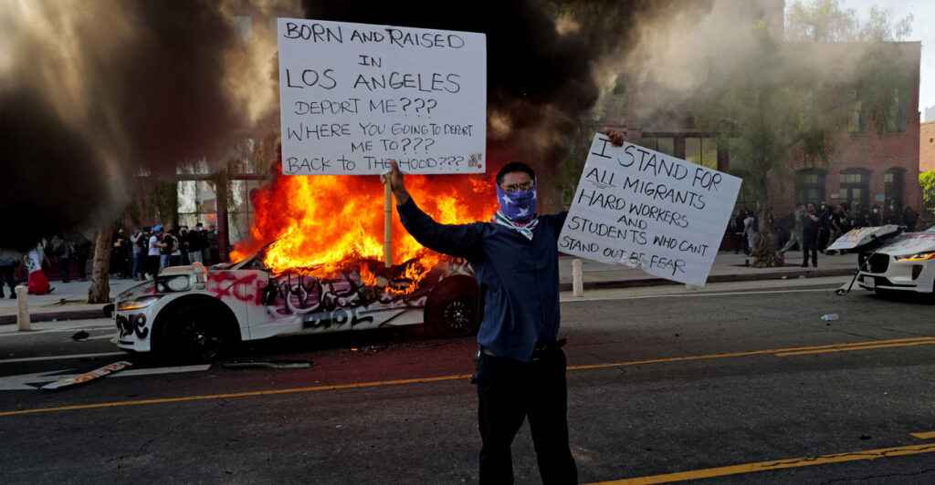 Masked rioter holds up signs protesting ICE deportations as car behind him is engulfed in flames filling sky with smoke.