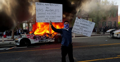 Masked rioter holds up signs protesting ICE deportations as car behind him is engulfed in flames filling sky with smoke.