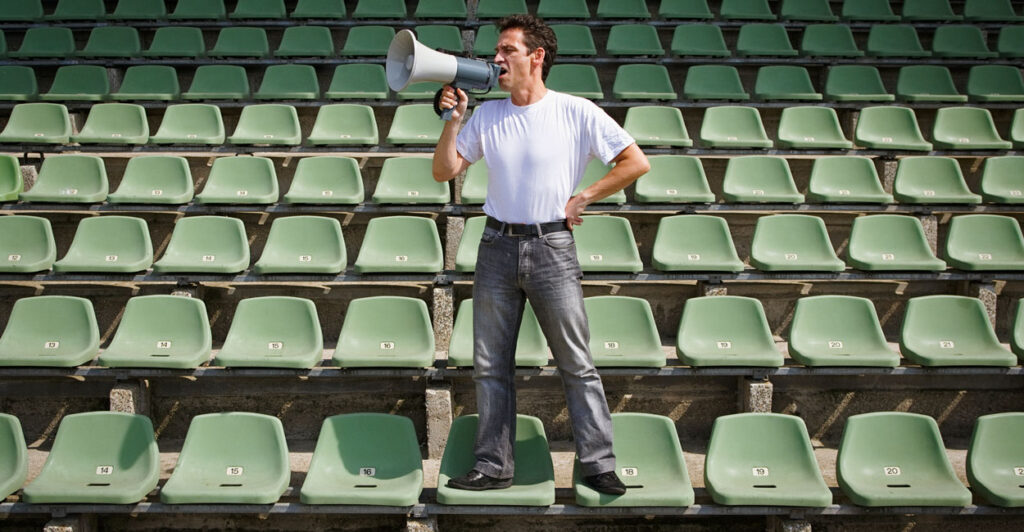 A man in a white shirt and jeans stands on seats surrounded by empty seats, shouting into a megaphone.