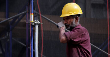 A bearded construction worker in yellow hard hat and burgundy shirt rests against a horizontal pole.