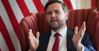 Close-up of Vice President JD Vance, sitting in a red chair, his hands raised, against a background of an American flag.