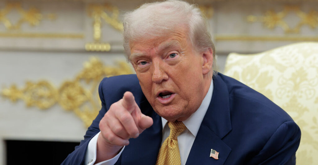 President Trump in blue suit and gold tie points at camera during Oval Office meeting.