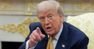 President Trump in blue suit and gold tie points at camera during Oval Office meeting.
