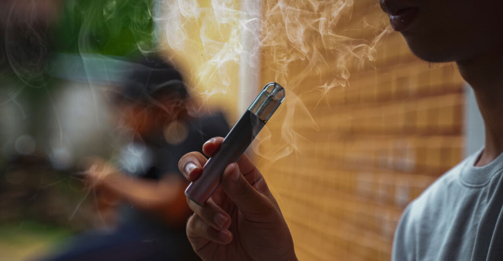 A young man vapes while leaning against a brick wall, smoke circling around his face.
