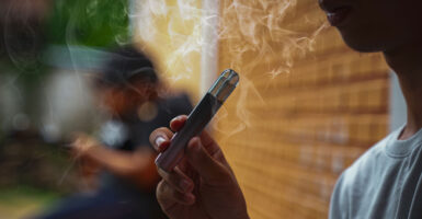 A young man vapes while leaning against a brick wall, smoke circling around his face.