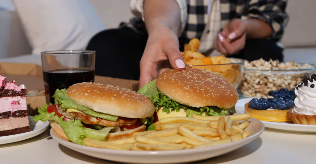 Woman reaches for a burger in the midst of a table of junk food.