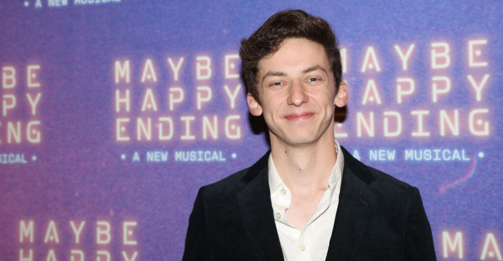 A smiling Andrew Barth Feldman stans in front of purple backing that reads "Maybe Happy Ending: A New Musical."