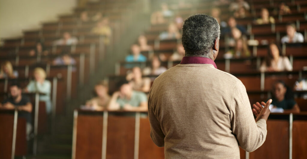 A black professor in tan sweater, with back to camera faces a lecture hall filled with students.