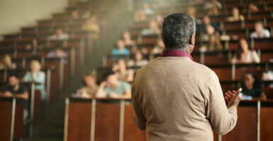 A black professor in tan sweater, with back to camera faces a lecture hall filled with students.