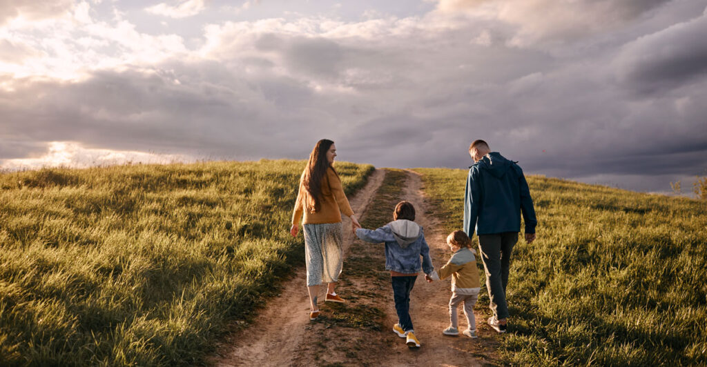 A mom and dad walk their two young children down a dirt road on an open plain, with a grand mostly cloudy sky ahead.