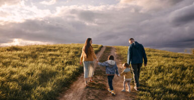 A mom and dad walk their two young children down a dirt road on an open plain, with a grand mostly cloudy sky ahead.