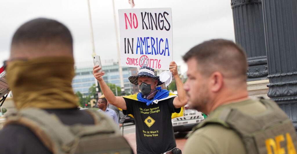 Aged protester in mask and black climate change shirt holds "No Kings in America" sign up in front of two DEA agents in Washington.