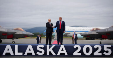 President Donald Trump and Russian President Vladimir Putin shake hands on a stage that says 'ALASKA 2025' on the front of it. Air Force One and fighter jets sit in the background.