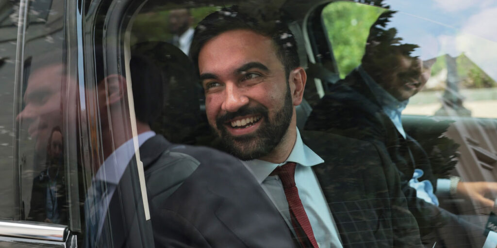 Zohran Mamdani smiles as he looks out the window of a car.