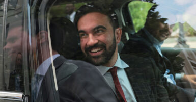 Zohran Mamdani smiles as he looks out the window of a car.
