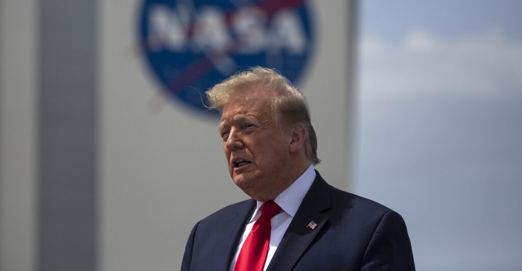 side view of Donald Trump in a blue suit and red tie speaking outside of a NASA facility with the NASA logo in back of him