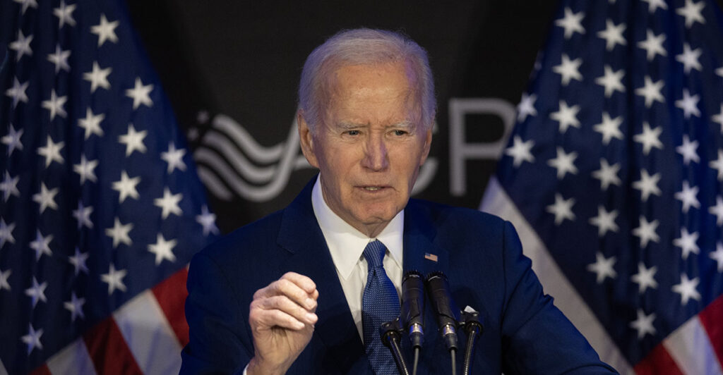 President Joe Biden in a blue suit gestures in front of American flags.