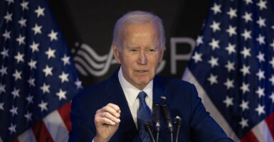 President Joe Biden in a blue suit gestures in front of American flags.
