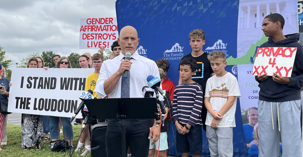 Seth Wolfe speaks in front of signs reading "I Stand With The Loudoun 3" and "Gender Affirmation Destroys Lives."