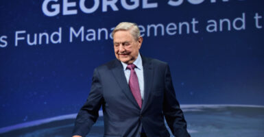 George Soros in a blue suit with purple tie against a blue background.