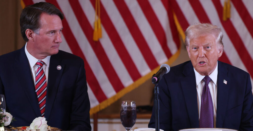 Governor Glenn Youngkin sits at a table next to Donald Trump. American flags in the background.