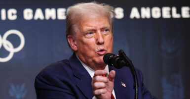 President Trump speaks to the press in front of a blue backdrop with white lettering reading "Olympic Games Los Angeles"
