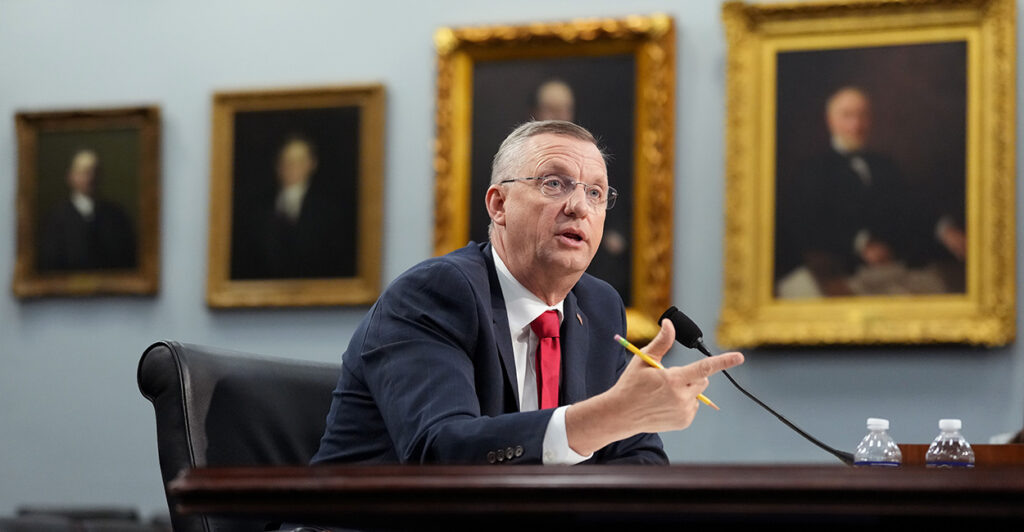 Veterans Affairs Secretary Doug Collins, seated at a desk with a microphone, and paintings on the wall behind him