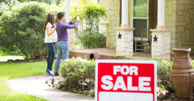 A couple stands in front of the house with a for-sale sign