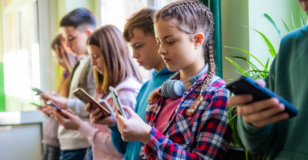 Young students are standing in the school hallway, all looking at their smartphones.