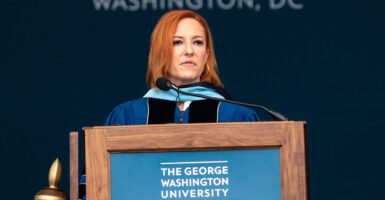 Jen Psaki wears a graduation gown as she stands behind a podium with a microphone as she looks out.