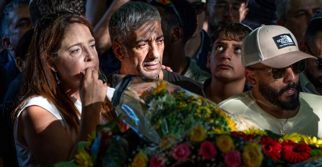A group of people mourn in front of wreath of flowers.