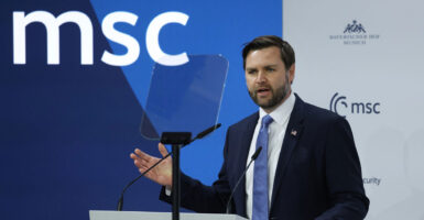 JD Vance sants before a podium and a microphone and speaks while holding out his right hand.