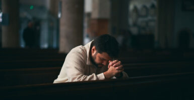 A man kneels in a church with his hands clasped in front of him as he puts his head down in prayer.