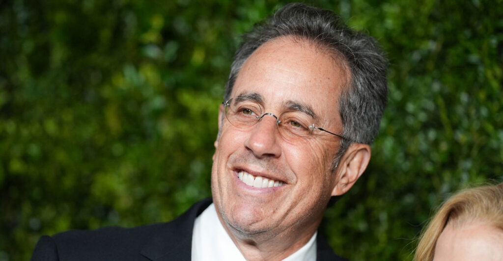 Jerry Seinfeld smiles while standing on the red carpet and looks to his left.