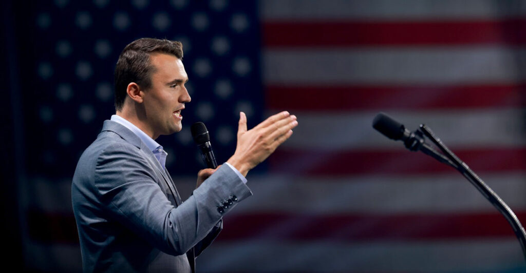 Charlie Kirk stands and holds a microphone in his left hand while gesturing with his right hand as he talks.