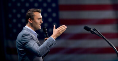 Charlie Kirk stands and holds a microphone in his left hand while gesturing with his right hand as he talks.